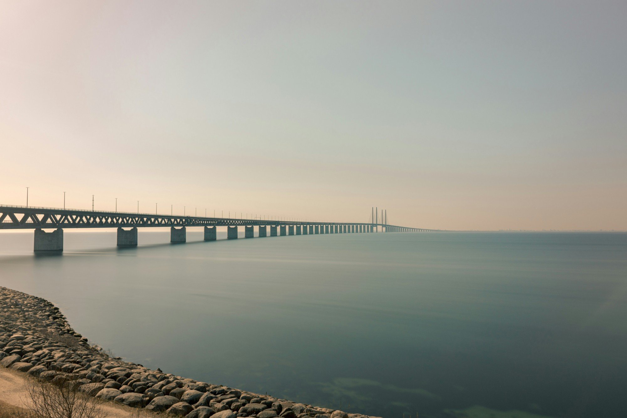 Öresundsbron sträcker sig ut över ett stilla Öresund i diset, sedd från stenstranden i Malmö