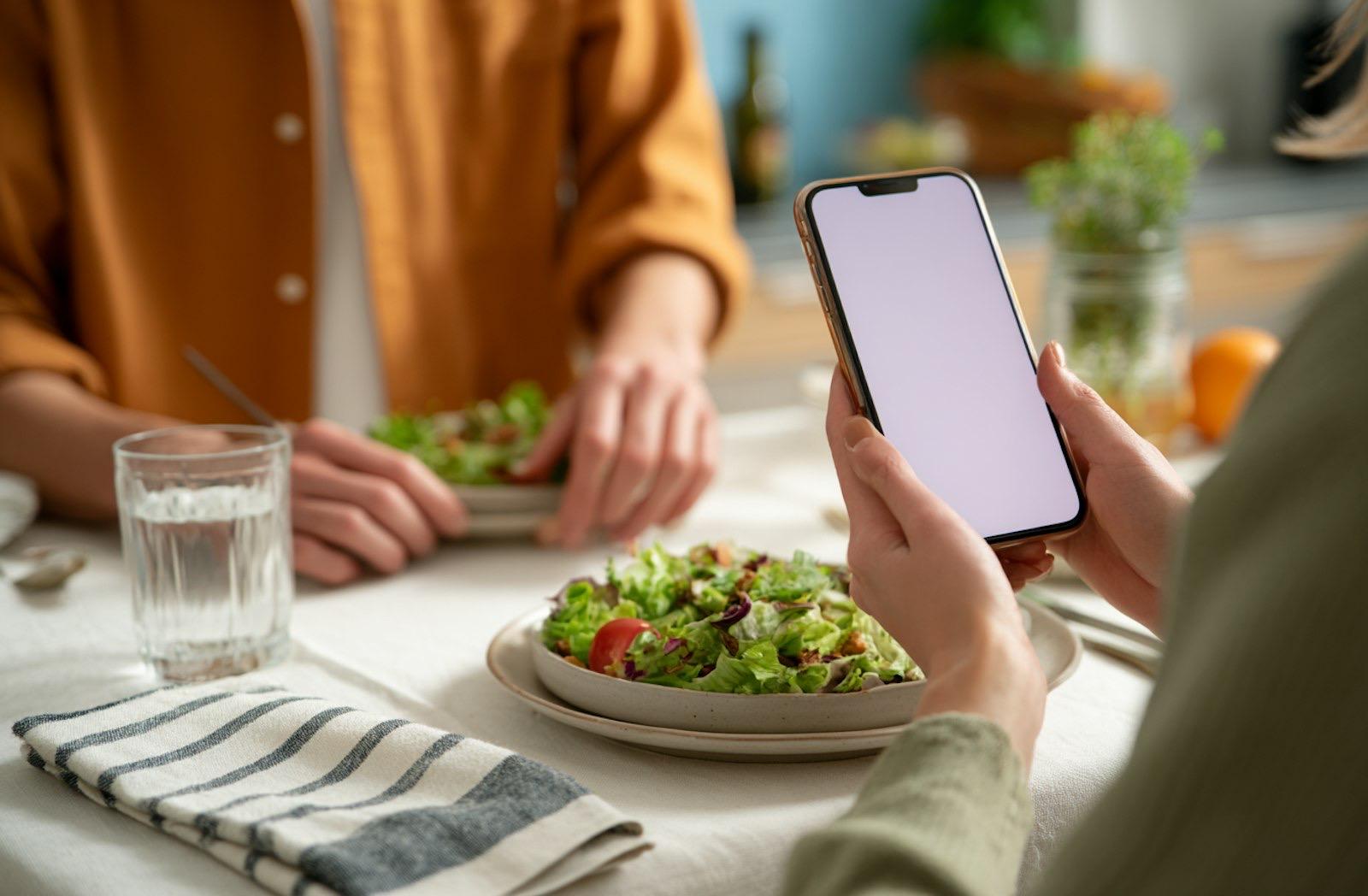 Mani che tengono uno smartphone a tavola, davanti a un piatto di insalata e a un bicchiere d’acqua, con un’altra persona seduta di fronte